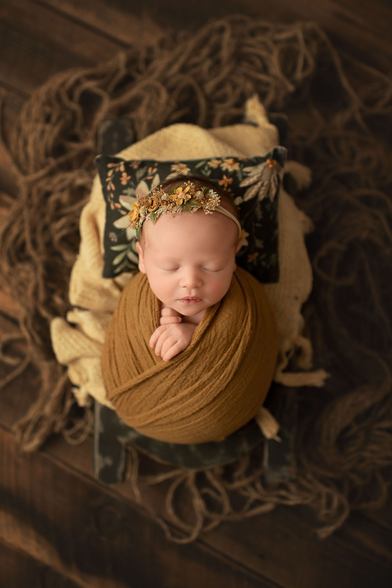Newborn wrapped in mustard fabric sleeping on floral pillow with decorative headpiece.