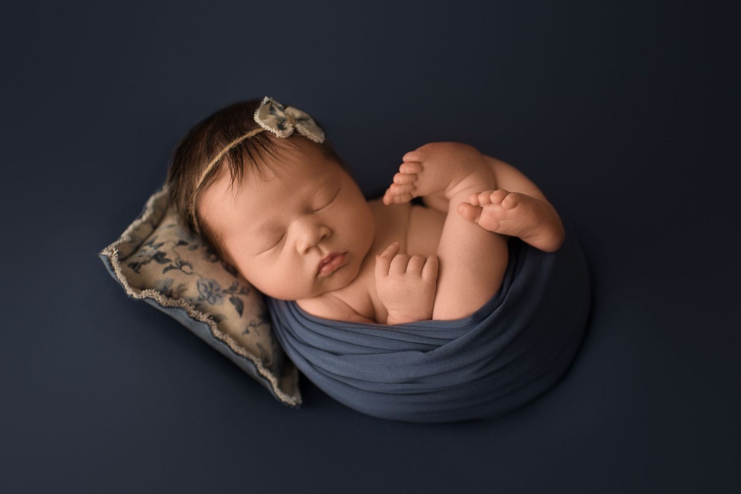 Sleeping newborn wrapped in blue fabric against dark background in artistic photography pose.