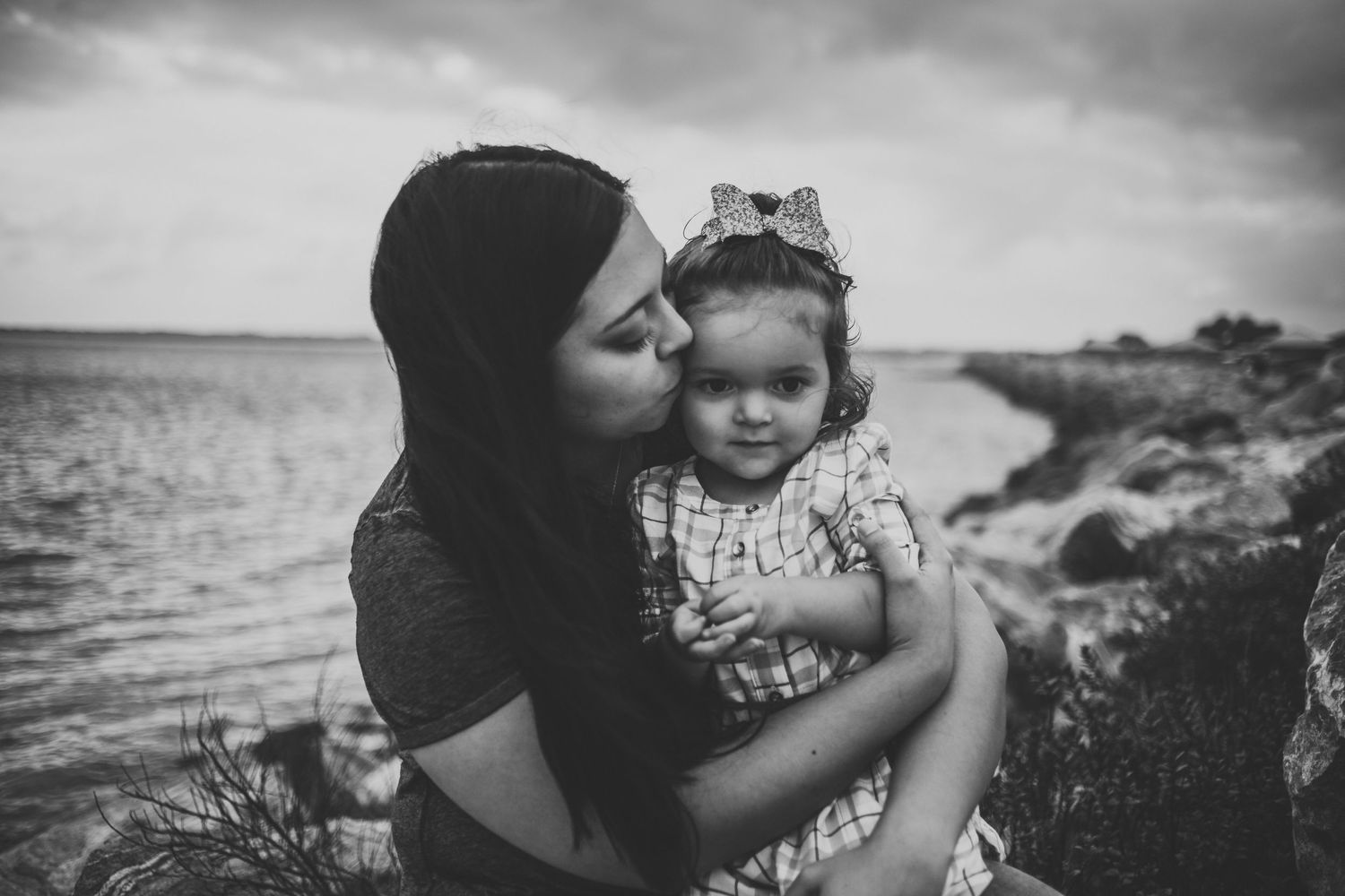 Black and white portrait of tender moment between parent and child near ocean shore.