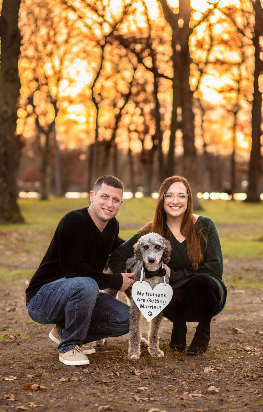A dog poses with a heart-shaped sign while a couple kneels beside them at sunset.