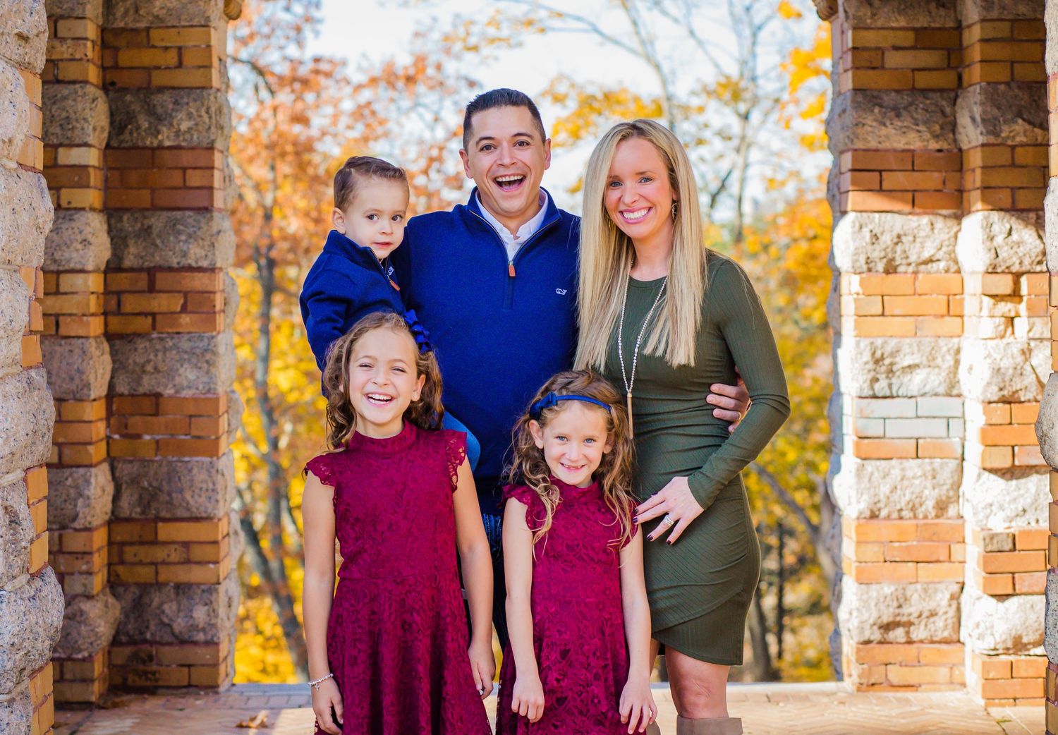 A family poses together in autumn between stone pillars with fall foliage in the background.