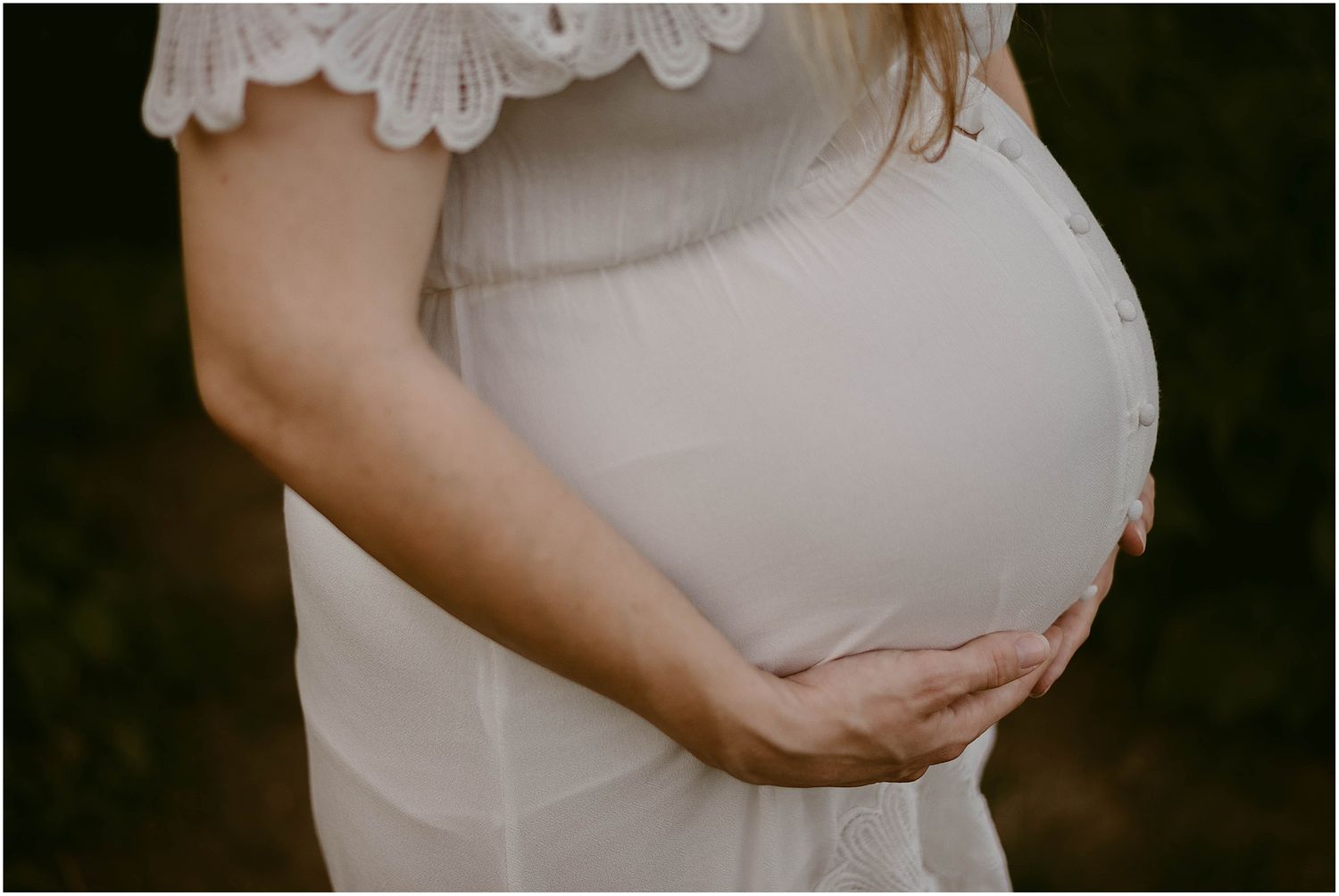 Close-up view of pregnant belly with hands tenderly cradling the bump in white dress.