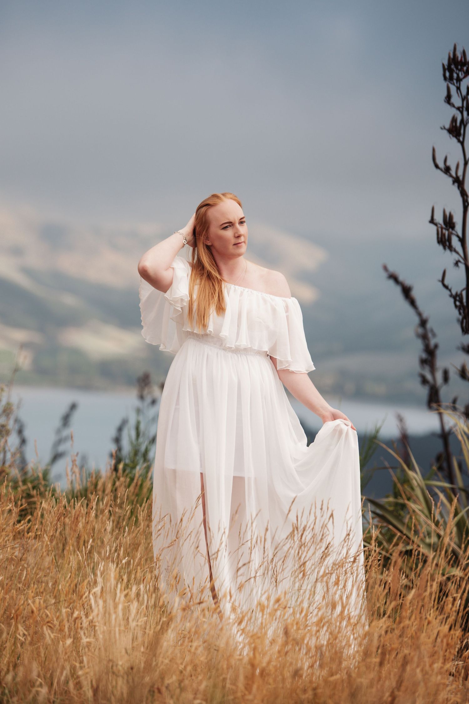 Ethereal portrait in flowing white dress standing in golden grass with mountains in background.