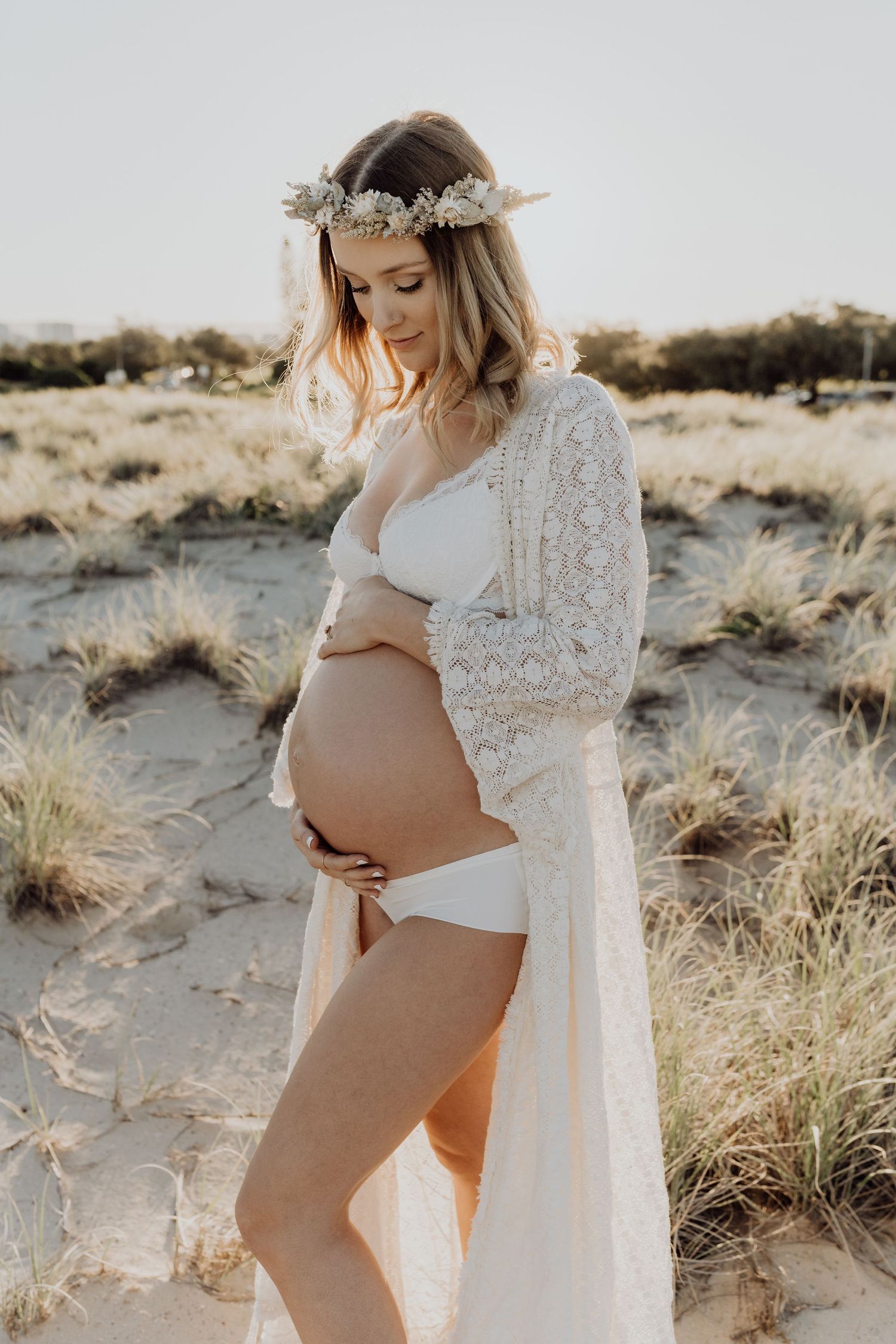 Ethereal maternity photo of an expectant mother in white lace robe and flower crown posing on sandy beach at sunset.