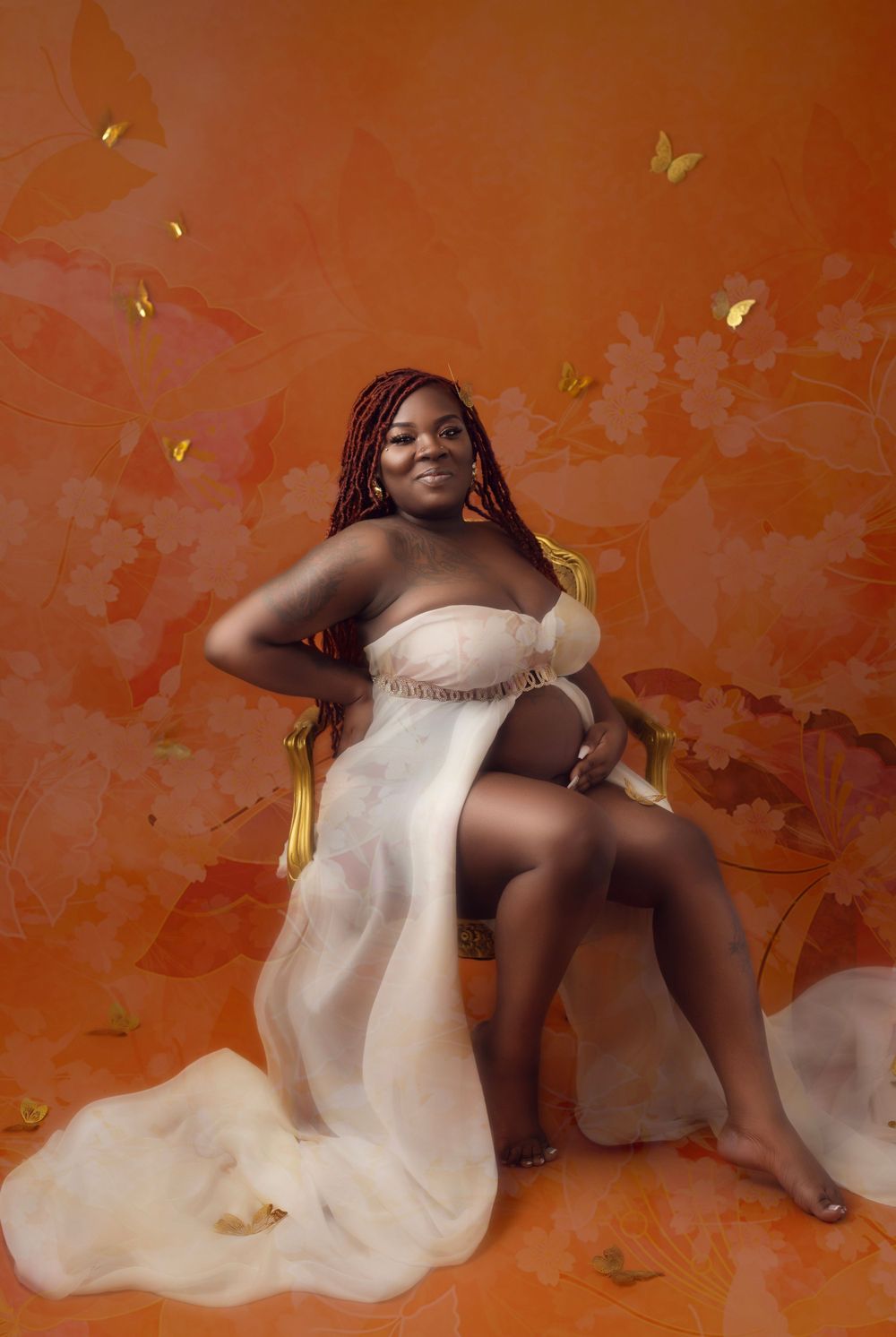 Artistic maternity photoshoot with flowing white fabric and gold petals against an orange backdrop.