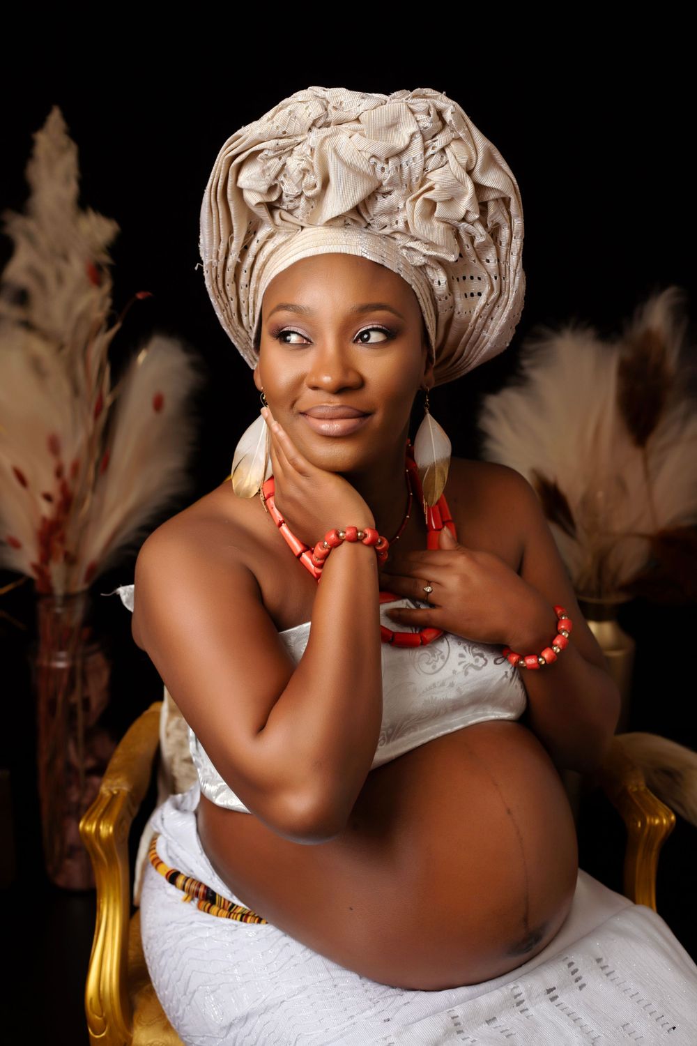 Maternity portrait with white dress, headwrap and coral accessories against dark backdrop.