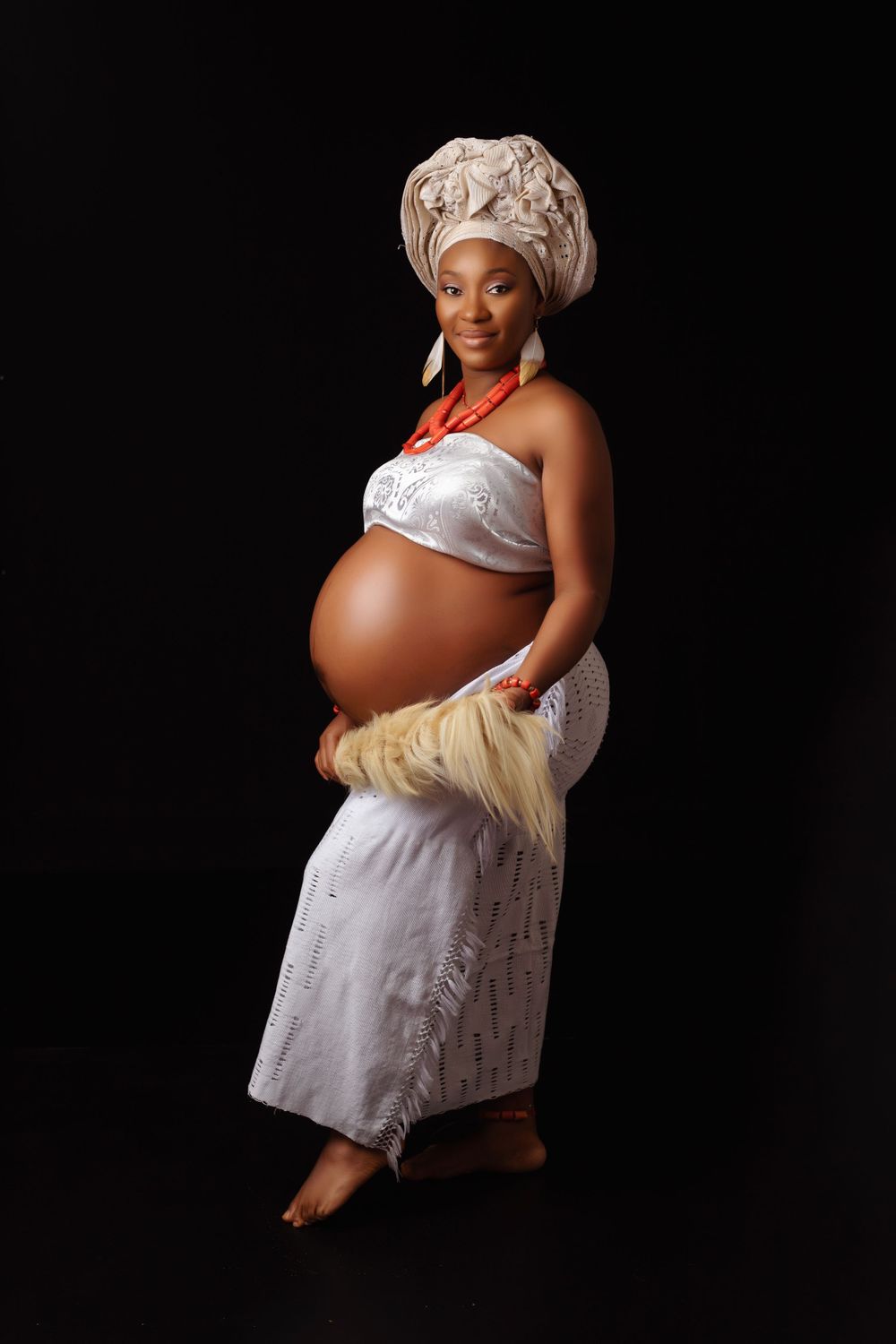 Elegant maternity portrait in white dress and headwrap against dark background.