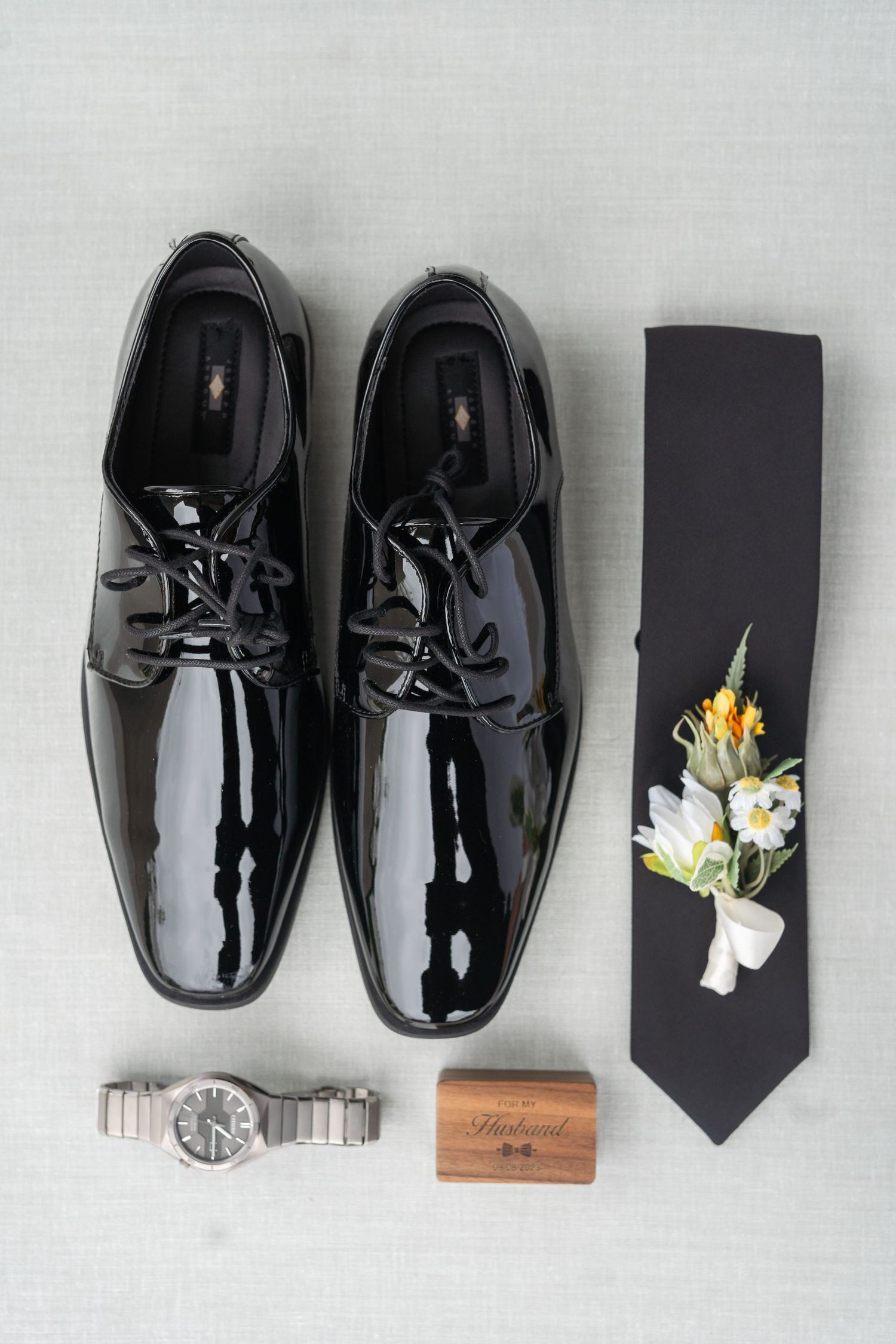 A pair of shiny black dress shoes, a boutonniere, and a tie are displayed on a white background.