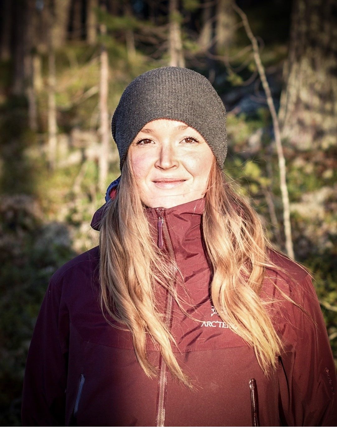 Smiling woman in a gray beanie and burgundy Arc'teryx jacket standing in a sunlit forest.