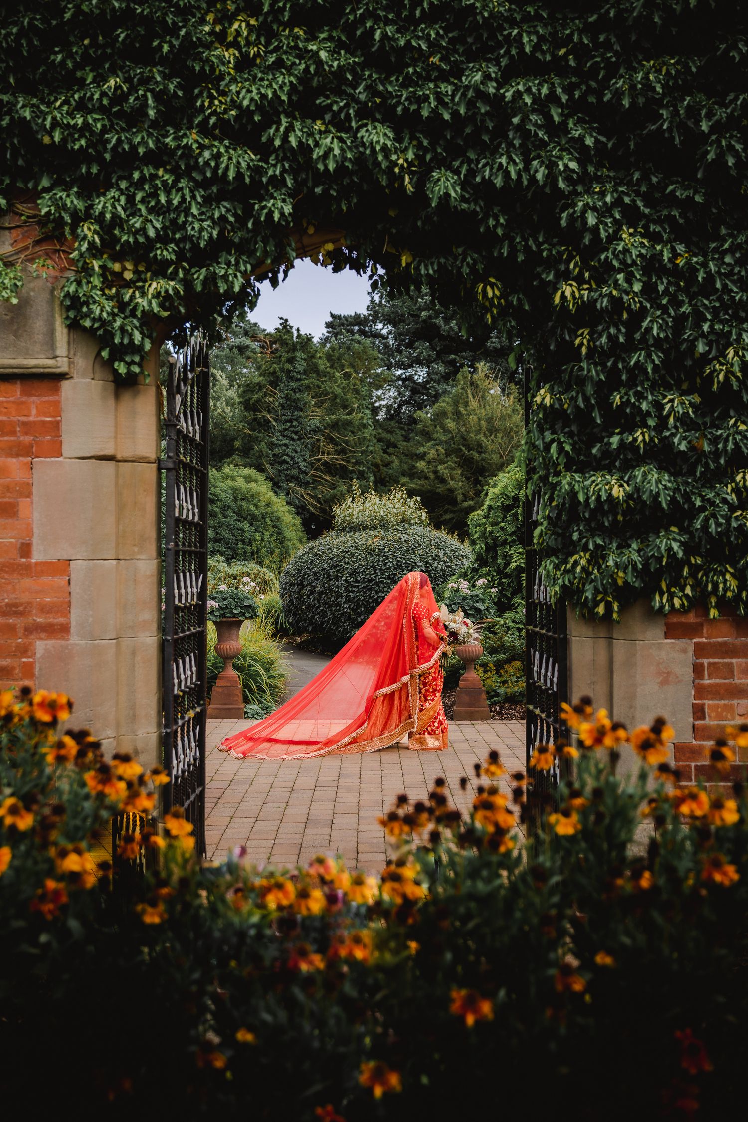 Vibrant red bridal dress trailing through an elegant garden archway framed by orange flowers and greenery.