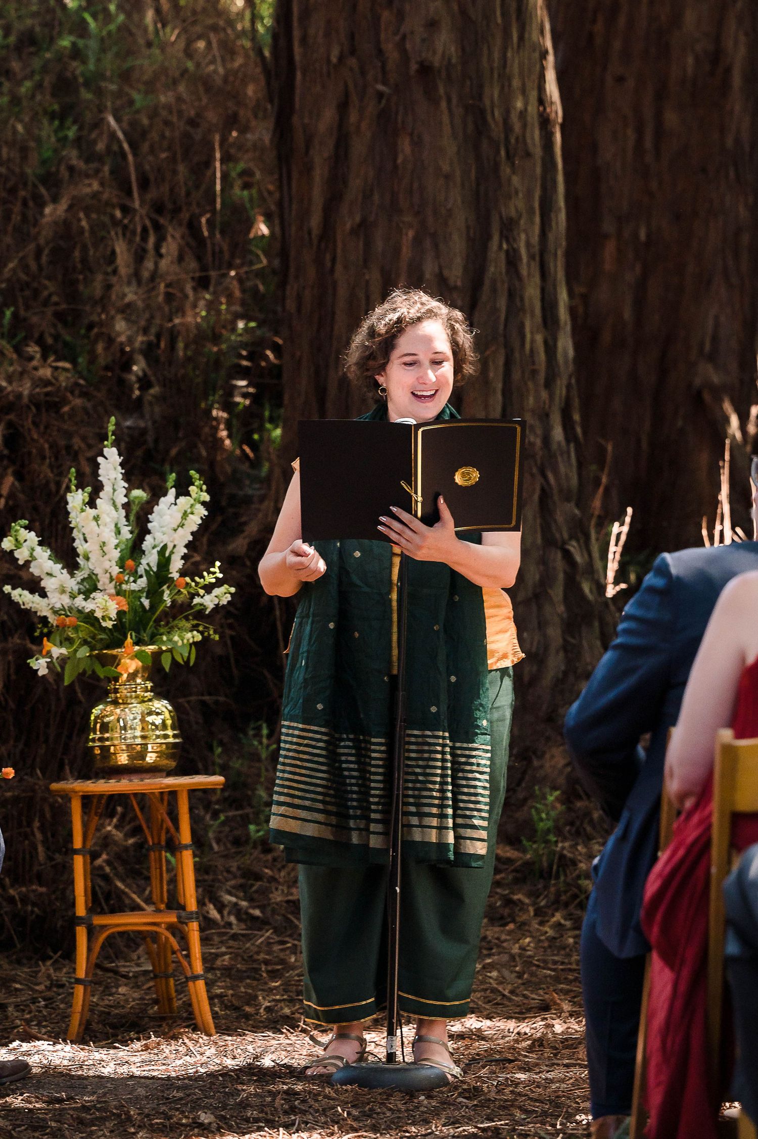 Wedding officiant in a dark green outfit holds a book during outdoor forest ceremony.