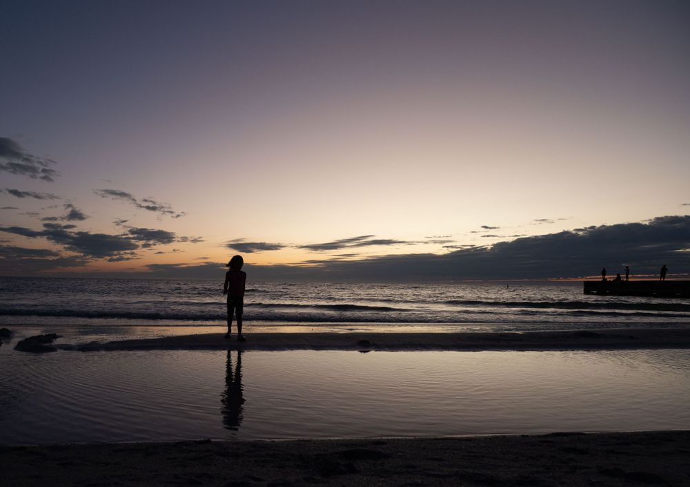 Silhouetted figure stands on reflective beach during dramatic sunset with dark clouds on horizon.