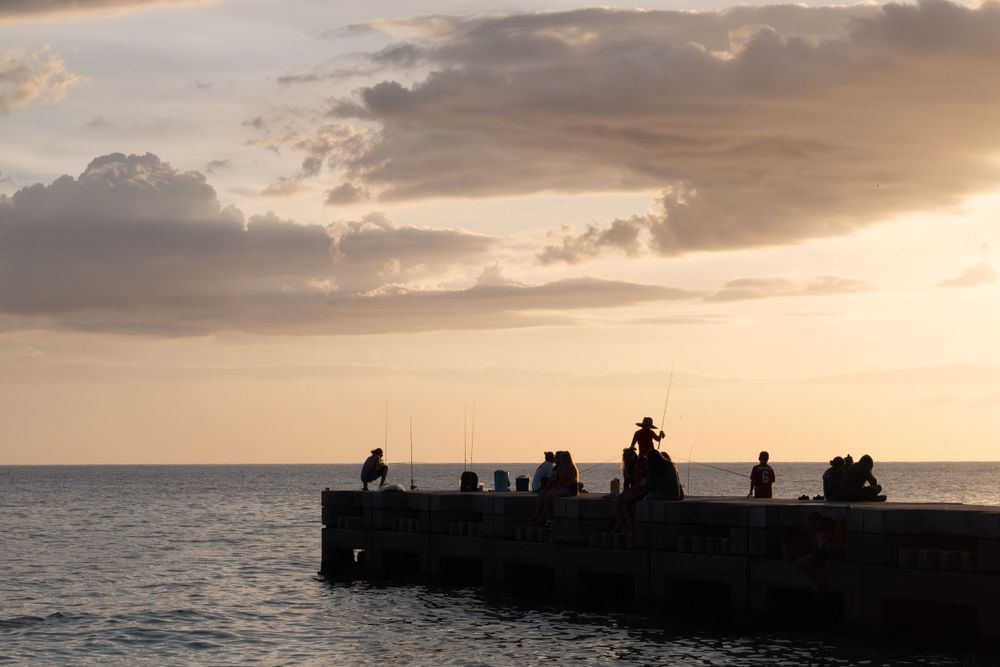People gather on pier silhouetted against dramatic ocean sunset with cloudy sky.