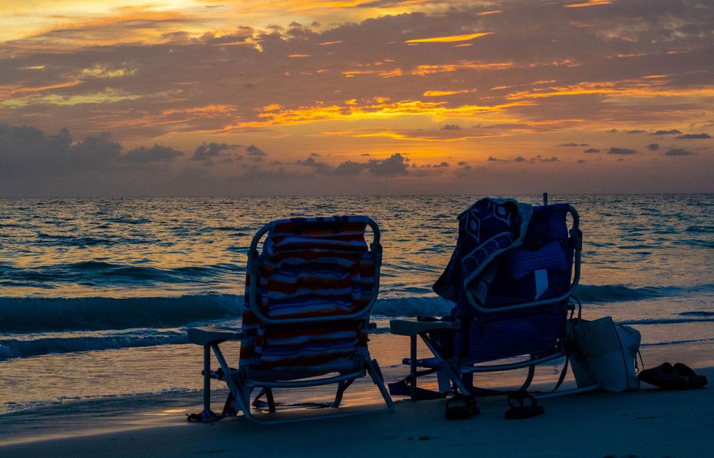 Two empty beach chairs silhouetted against vibrant orange sunset over ocean waves.