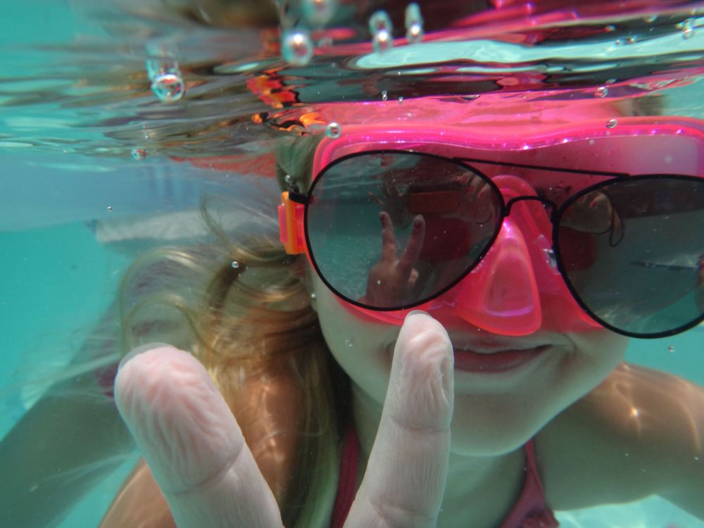 Underwater view of snorkeler wearing pink mask and sunglasses making peace sign.
