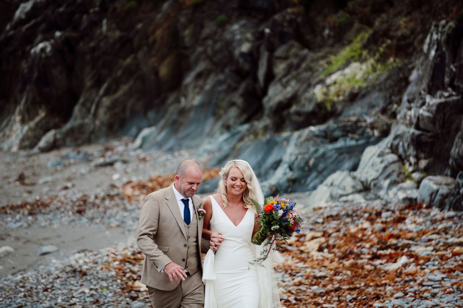 A wedding couple shares a joyful moment together on a rocky beach with cliffs behind them.