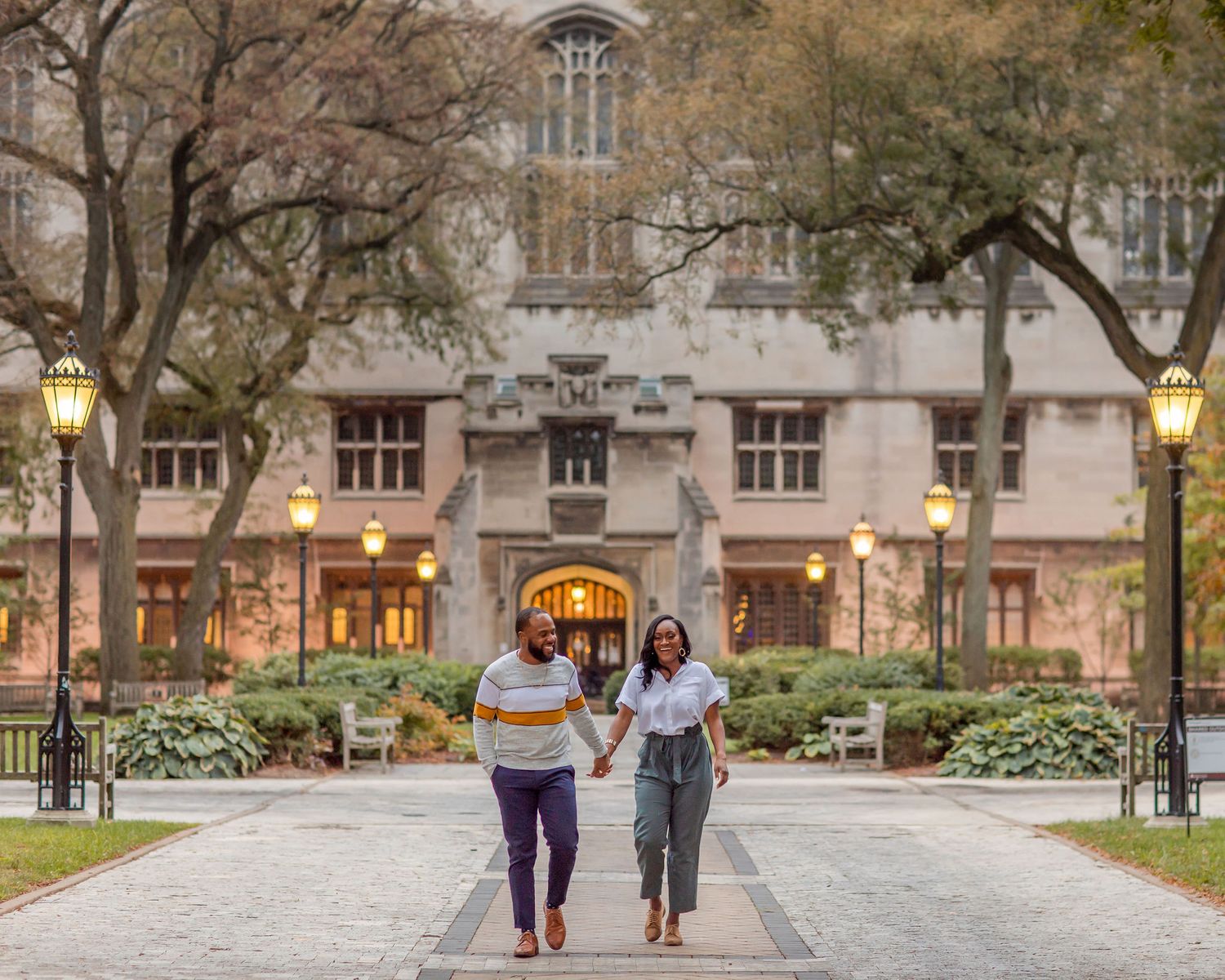 Two students walk along a tree-lined path at a university campus with Gothic architecture in the background.