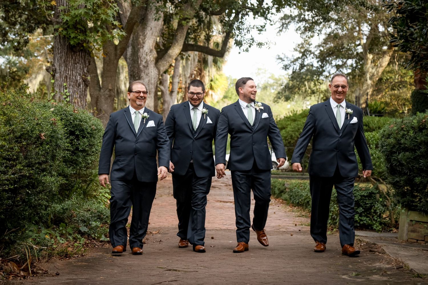 A group of groomsmen in matching black suits walk together along a tree-lined path.