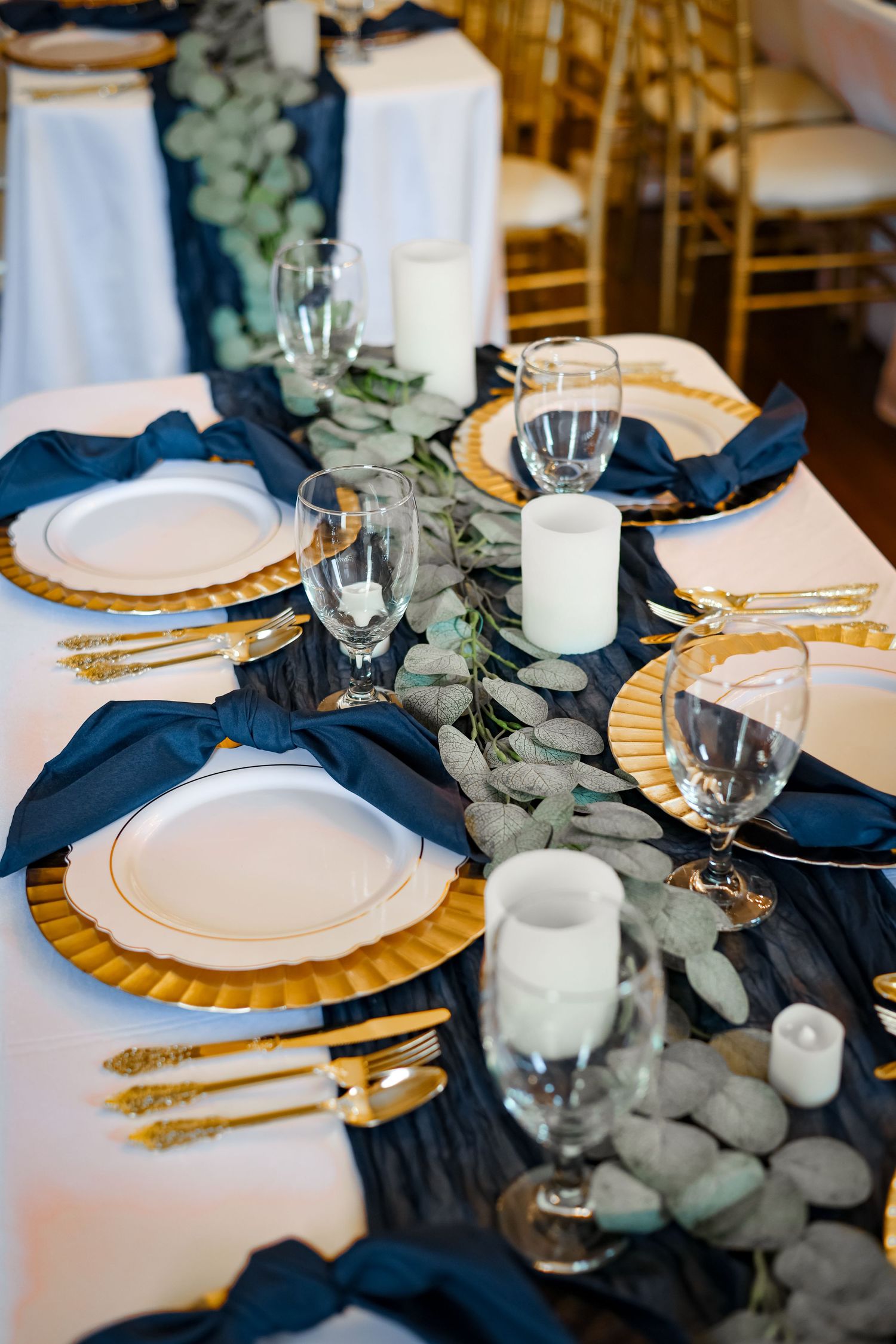 An elegant wedding table setting with navy linens, gold chargers, and eucalyptus centerpiece.