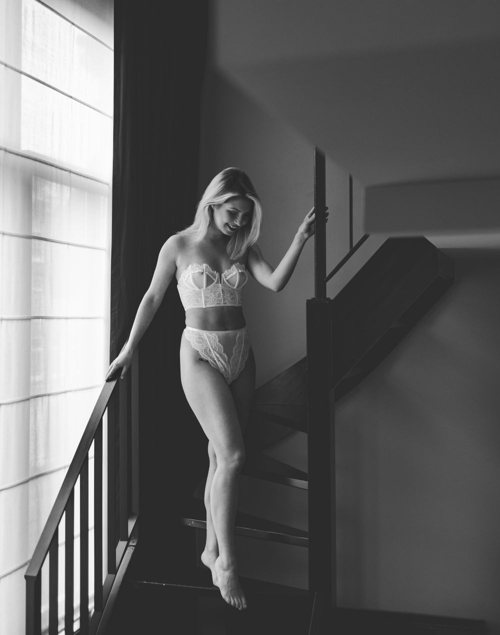 Artistic black and white portrait of figure posing on stairs near window.
