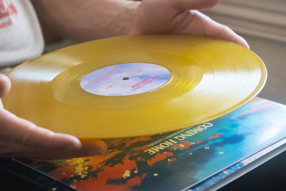 Hands holding yellow vinyl record over colorful album artwork.