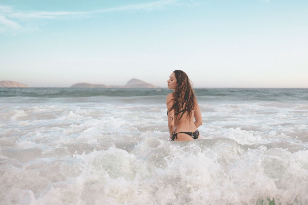 A person sits in ocean waves during sunset with mountains visible in the background.