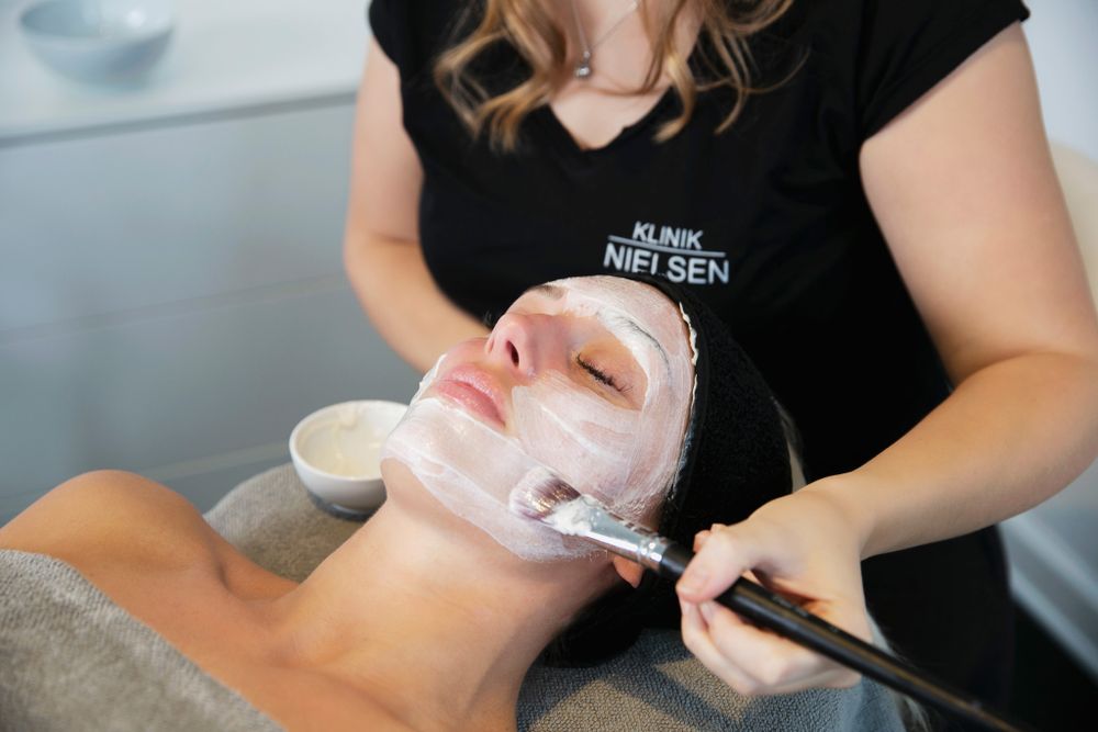 En beautyterapeut påfører forsigtigt en hvid ansigtsmaske under en spa-behandling.