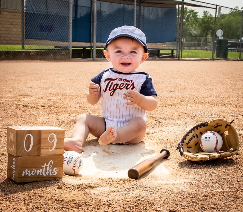 Baby in baseball outfit sitting on field with glove, bat and age milestone blocks showing 9 months.
