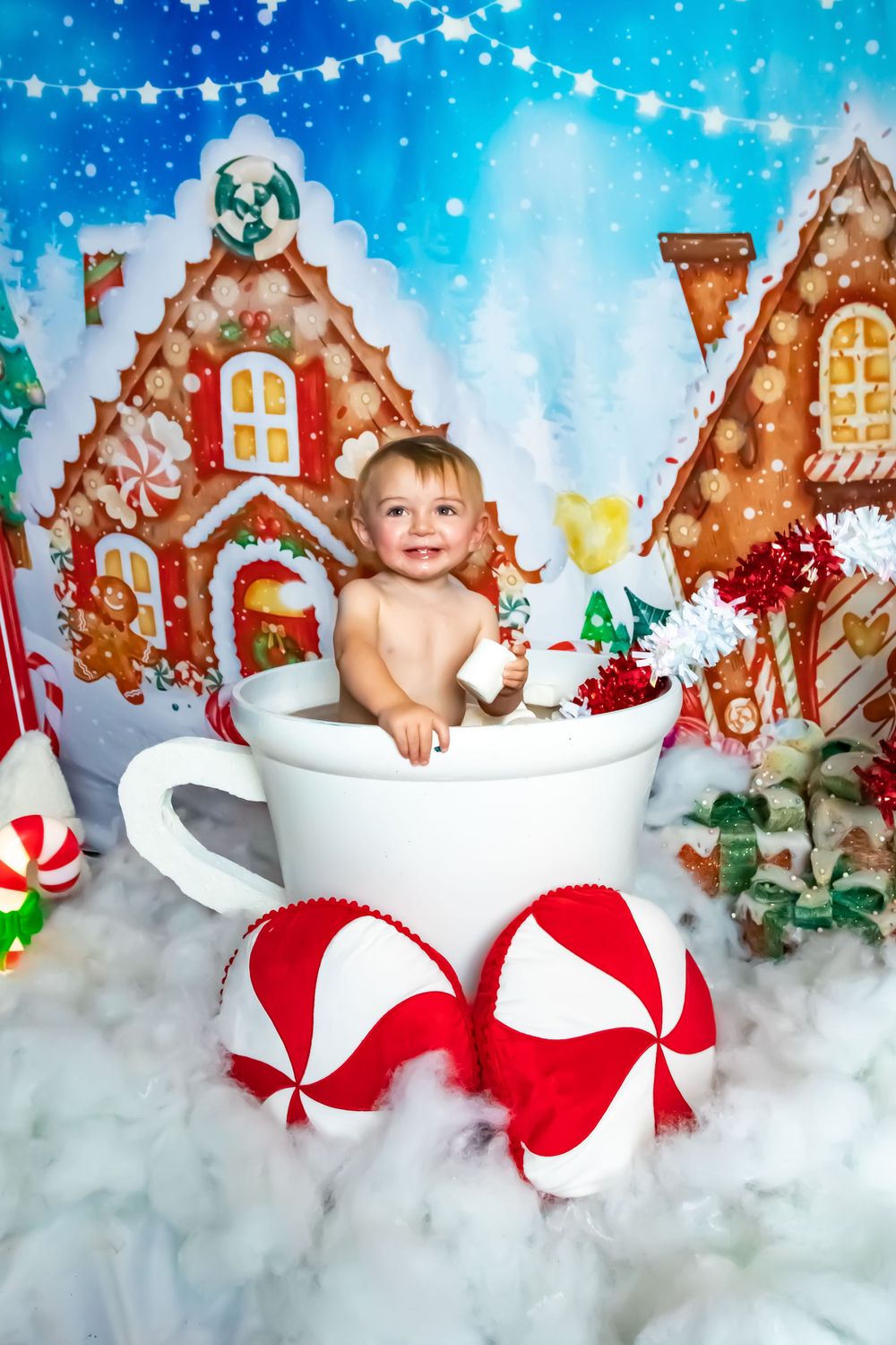 Baby sitting in oversized coffee cup prop with gingerbread house backdrop and peppermint decorations.