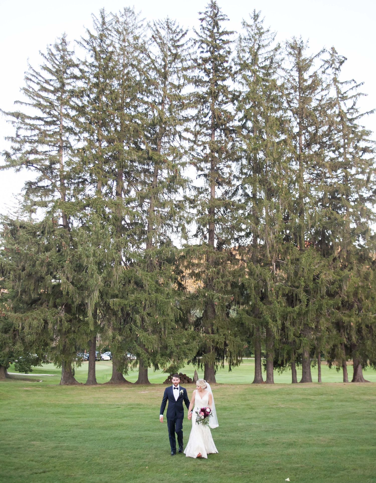 A newlywed couple walks across a lush green lawn beneath towering evergreen trees.