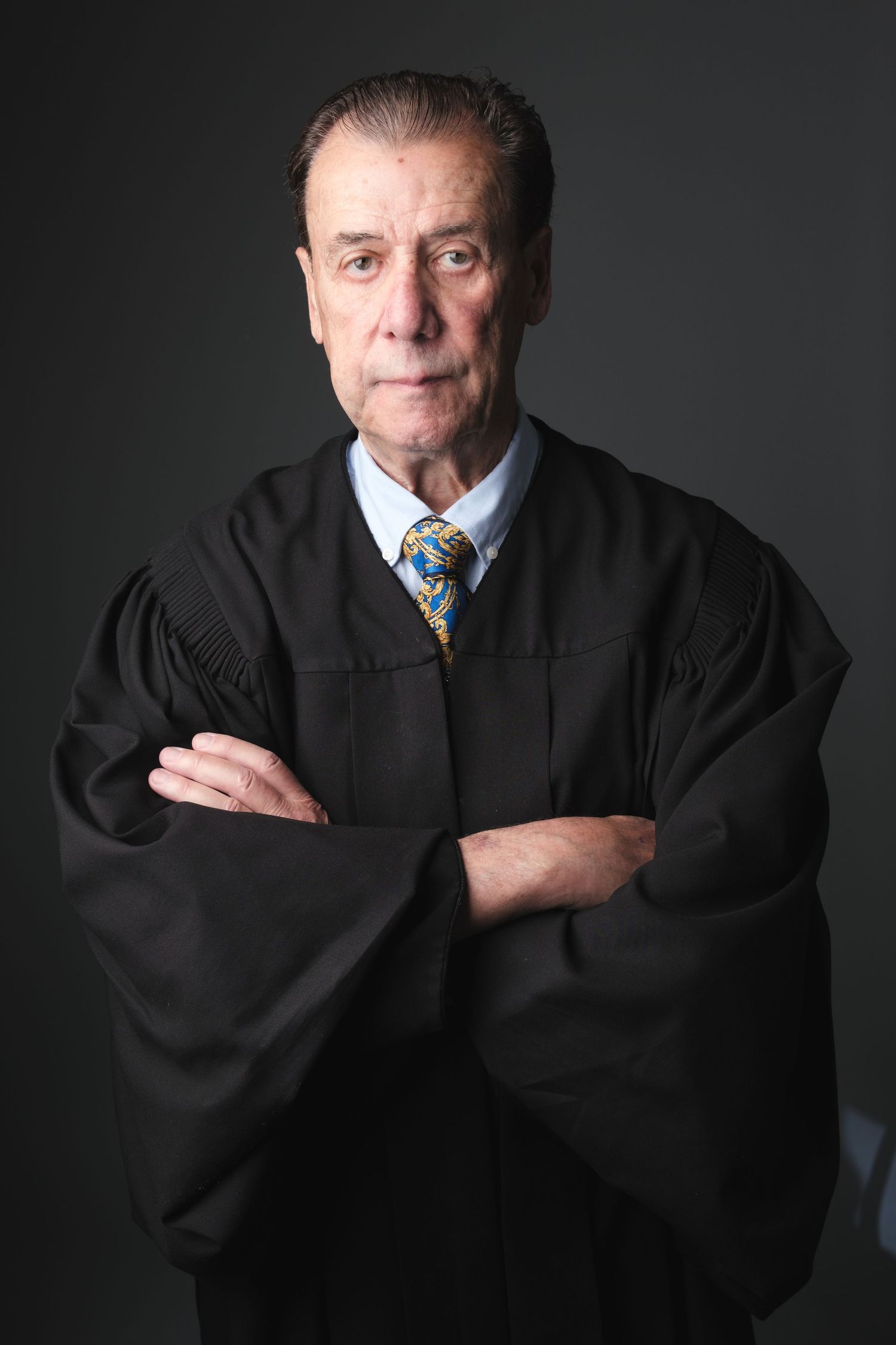 Professional portrait of a judge in traditional black robes and blue tie against a dark background with arms crossed.