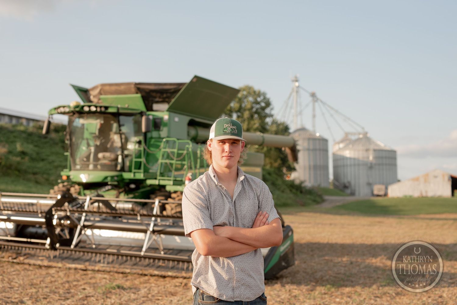 Farmer poses with grain silos and harvesting equipment in rural agricultural setting.