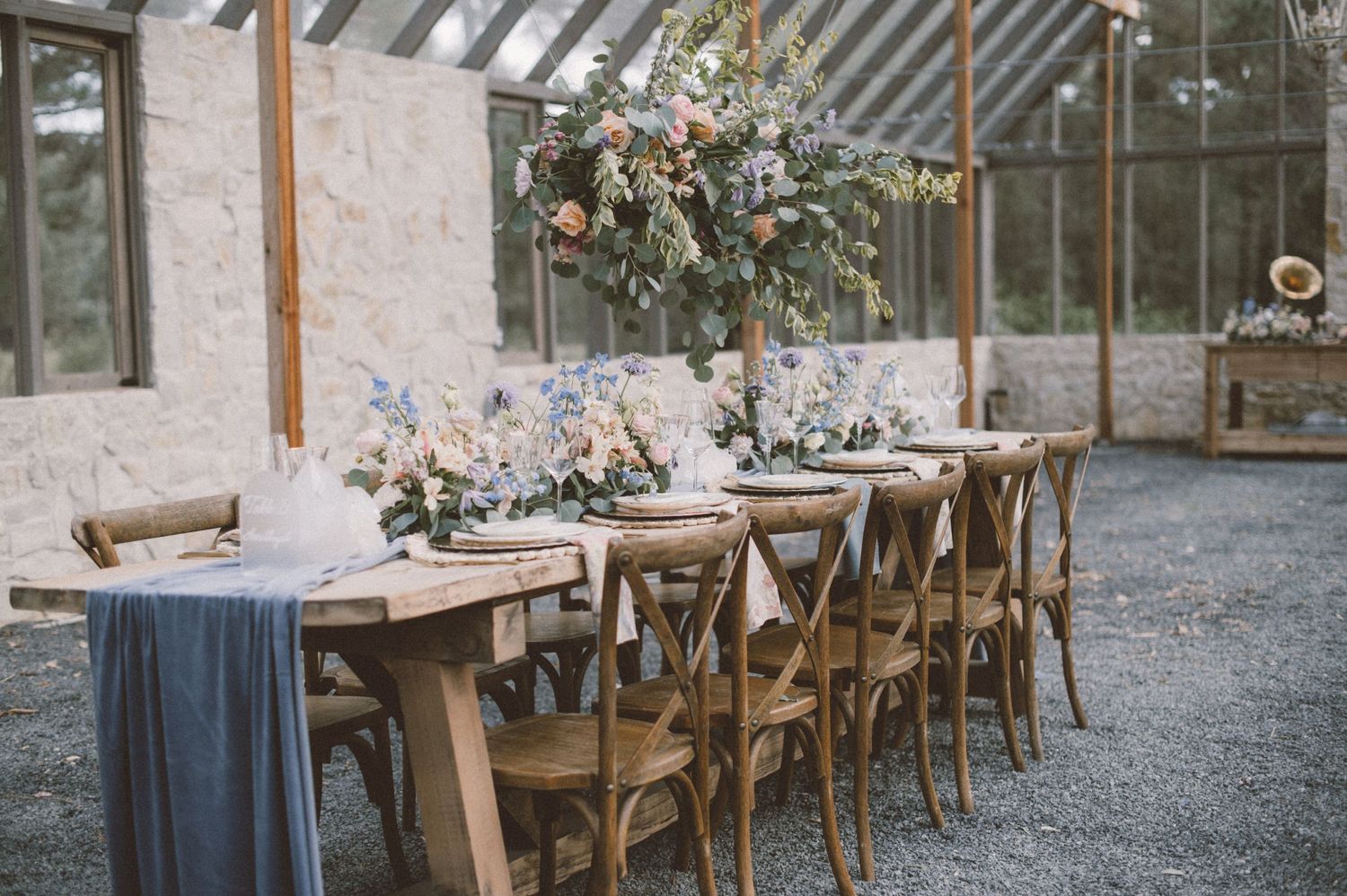 Rustic wooden dining table set with elegant floral centerpieces in a light-filled greenhouse venue.
