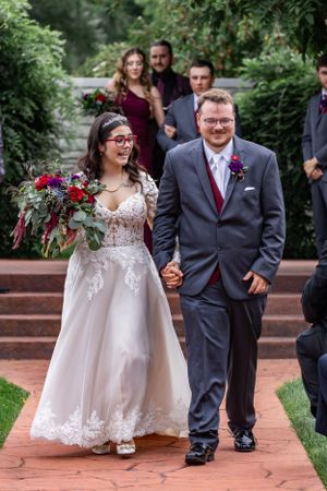 A happy wedding couple walking down outdoor brick steps surrounded by greenery and red flowers.
