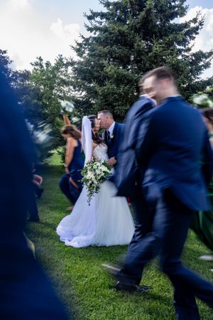 A wedding couple kisses while surrounded by guests in navy suits against an outdoor evergreen backdrop.