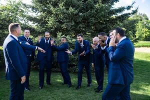 Groomsmen in navy blue suits share a laugh together on a grassy lawn near evergreen trees.