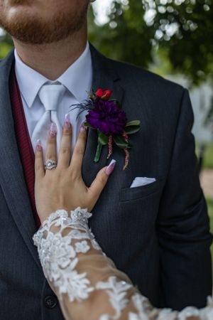 Close-up of a purple and red boutonniere on a dark suit with bride's lace sleeve and ring visible.
