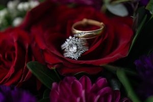 Diamond engagement ring and wedding band nestled on a vibrant red rose with purple flowers.