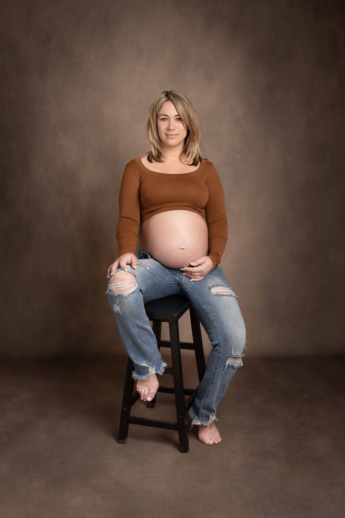 Maternity portrait of woman in brown sweater and distressed jeans sitting on wooden stool.