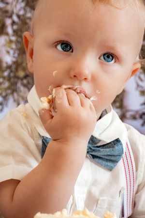 A toddler in a white shirt and blue bow tie enjoying cake during a birthday celebration.