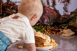 A child in white leaning over to taste a birthday cake on the floor.