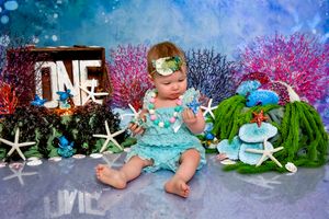 Baby in aqua dress sits among starfish decorations and pink flowers in an underwater themed photo setup.