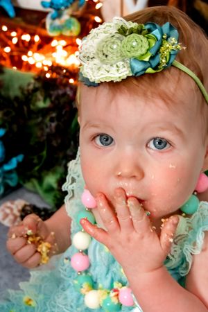 Baby wearing mint green dress and floral headpiece with twinkling lights in background.