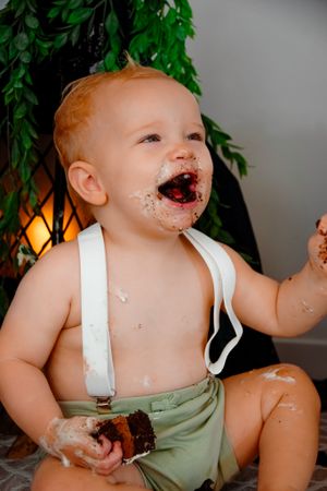 Baby in suspenders enjoying messy cake smash with chocolate frosting everywhere.