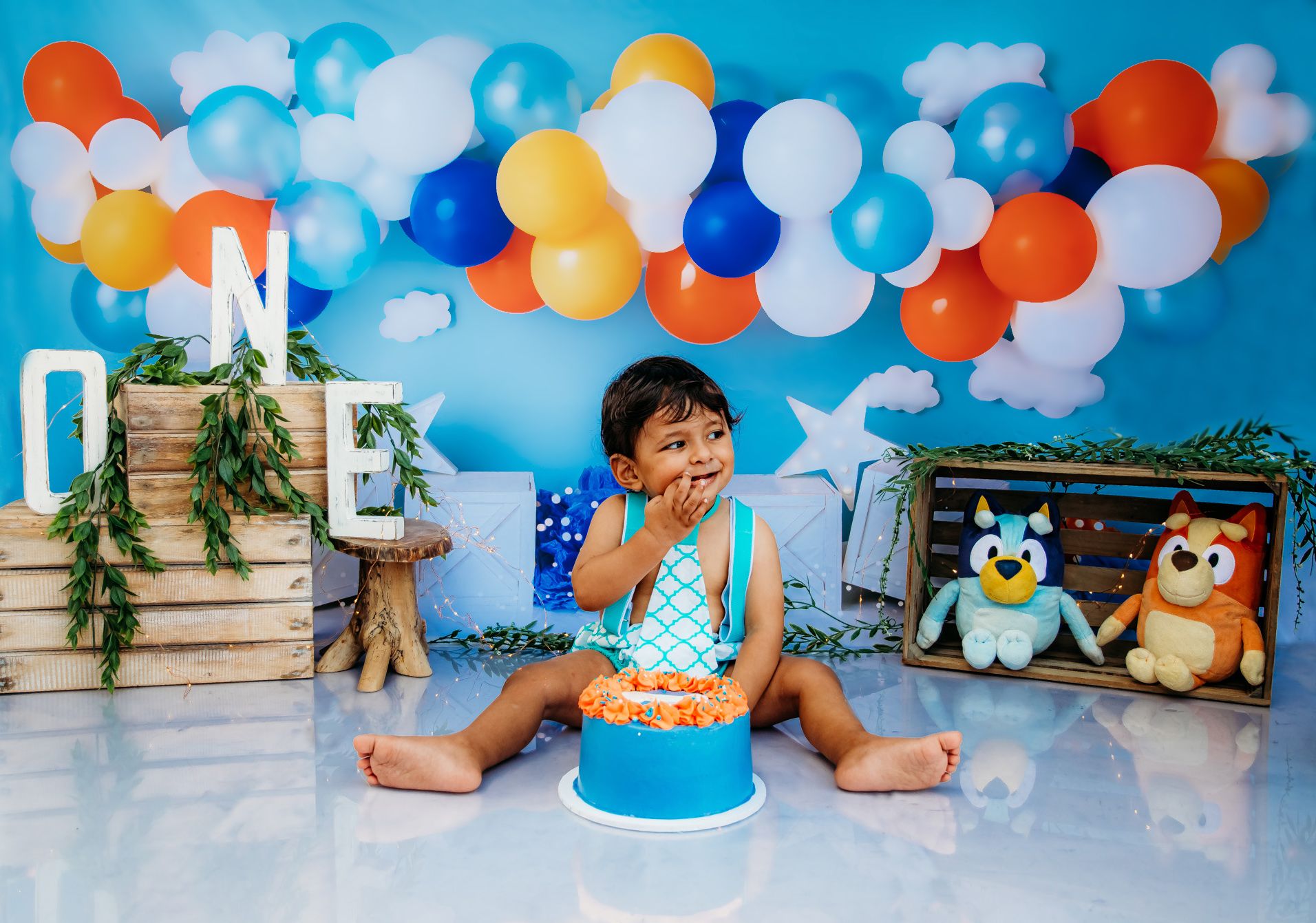 Festive first birthday setup with blue and orange balloons against a cloudy sky backdrop.