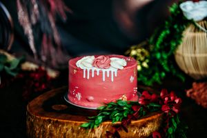 Red and gold wedding cake decorated with greenery on a wooden stand in dramatic lighting.