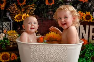 Two young children sitting in a white tub surrounded by sunflowers and pumpkins.