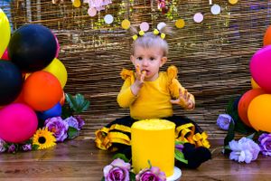 Baby in yellow outfit sitting with colorful balloons and cake during first birthday celebration.