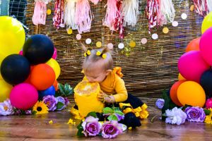 Baby in yellow outfit reaching for cake surrounded by balloons and flowers at birthday party.