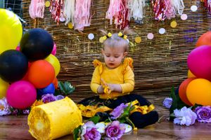 Baby sitting with smashed yellow cake surrounded by colorful party decorations.